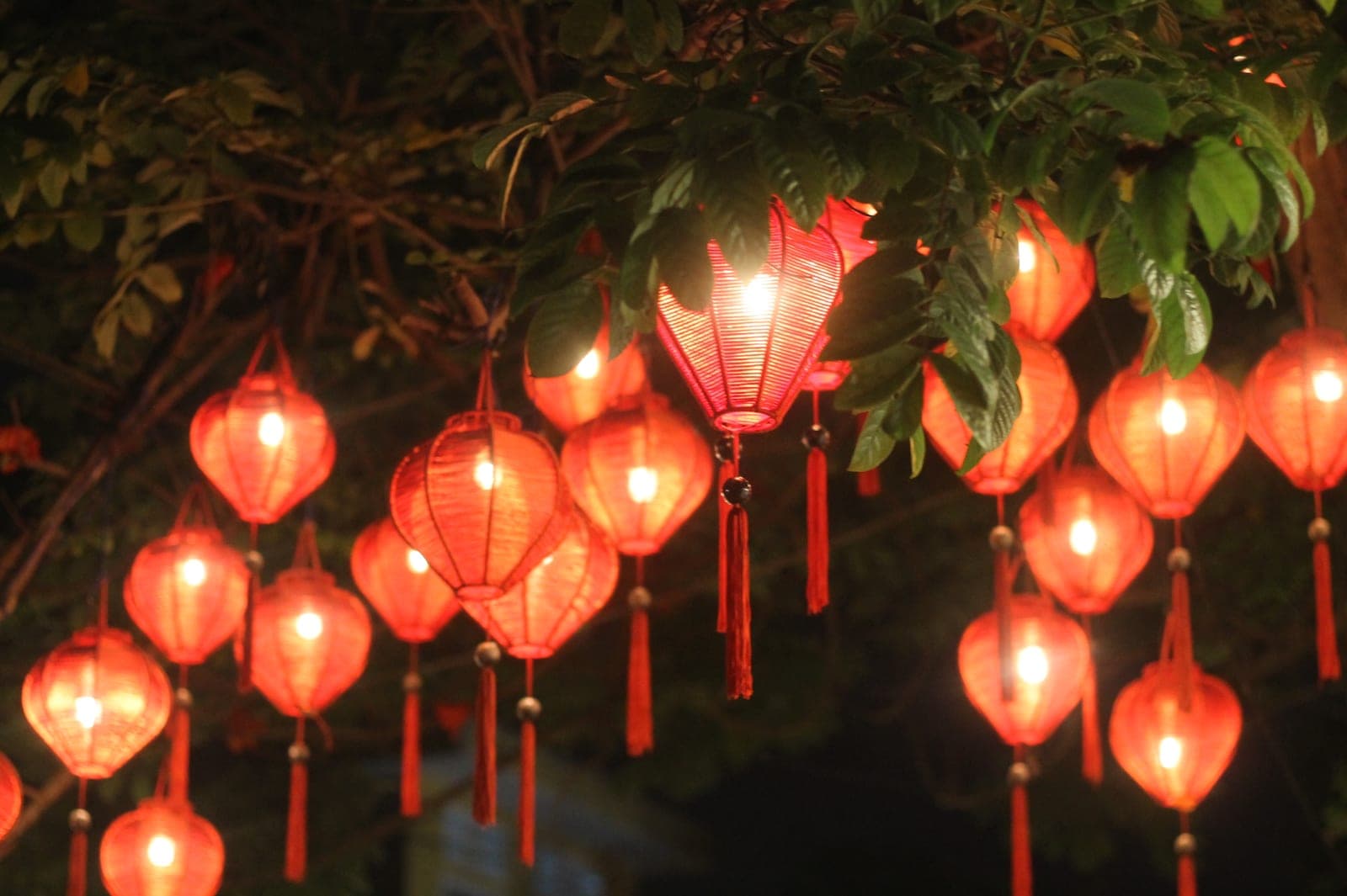 Glowing silk lanterns in warm colors hanging over Hoi An's Old Town streets at dusk, with reflected light on the water below.
