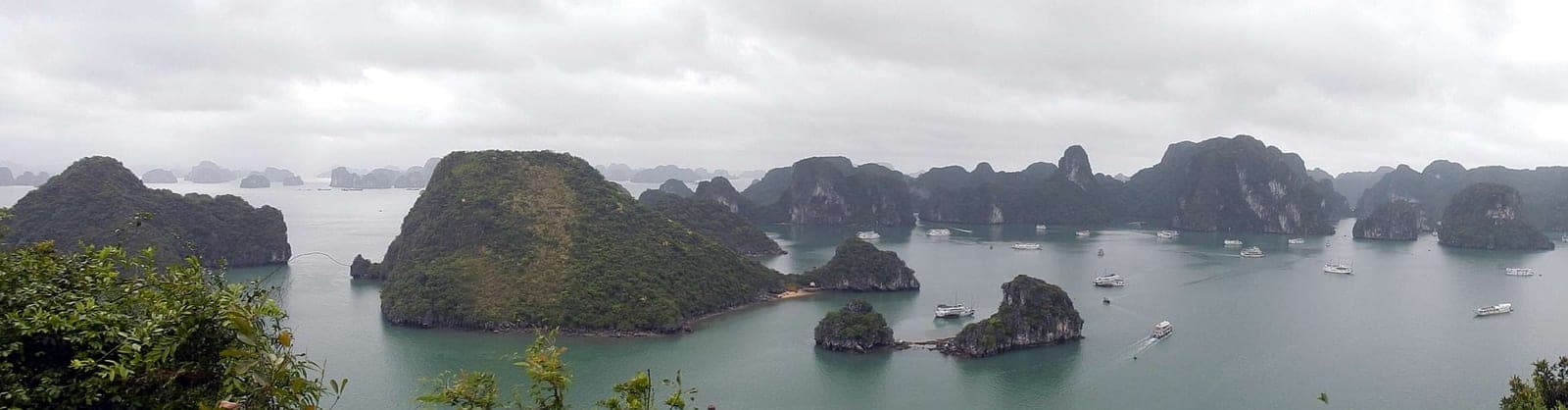 Sweeping panoramic view of Ha Long Bay's limestone karsts rising from emerald water under a light sky, Vietnam.