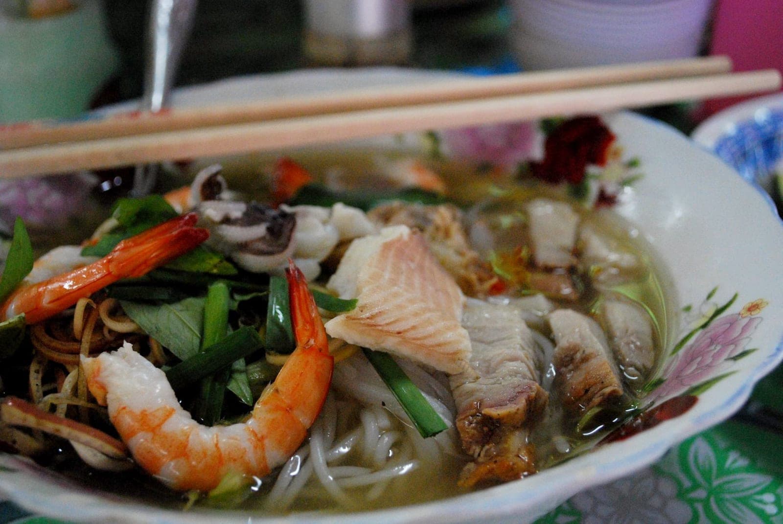 A bowl of bun mam with noodles, pork belly, prawns, squid, and dark fermented fish broth served at a market stall