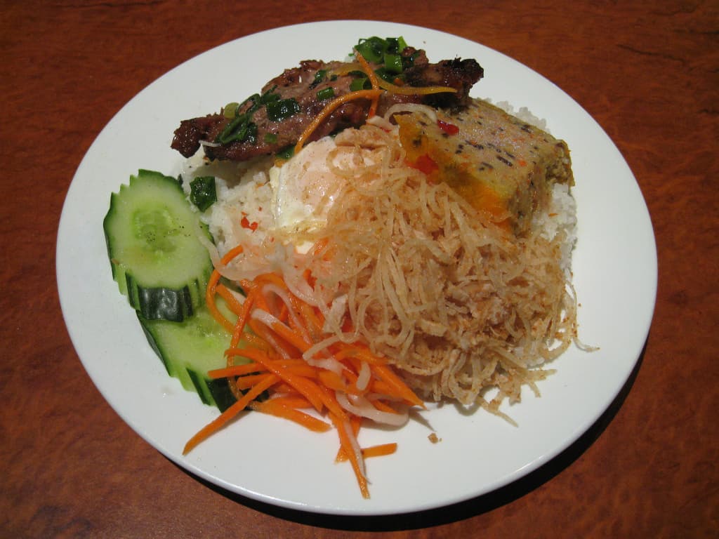 A plate of cơm tấm with grilled pork chop, fried egg, pickles, and broken jasmine rice on a white plate with small sauce bowl.