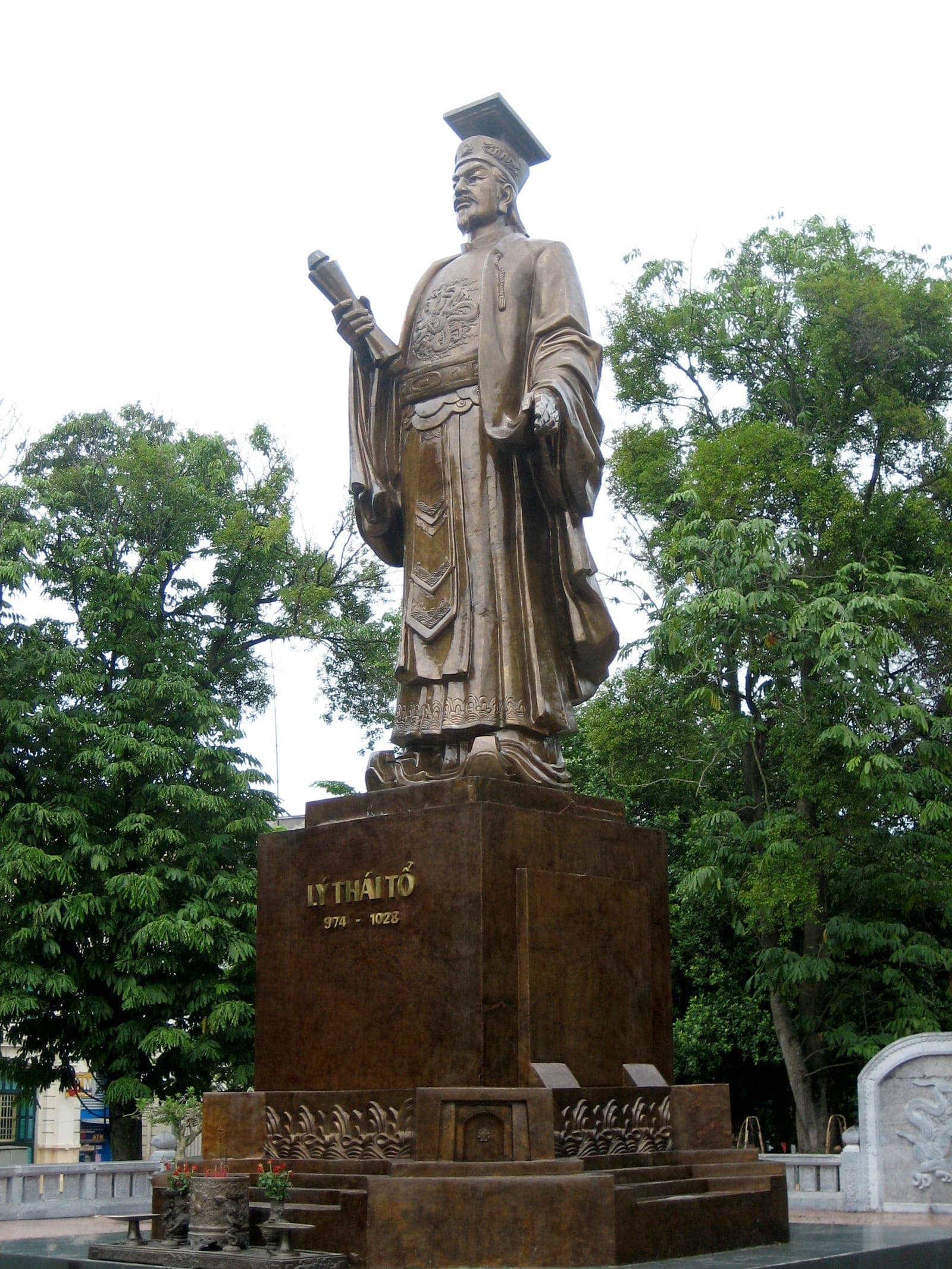 Bronze statue of a robed seated figure with one hand resting on a scroll, located in a park beside Hoàn Kiếm Lake.