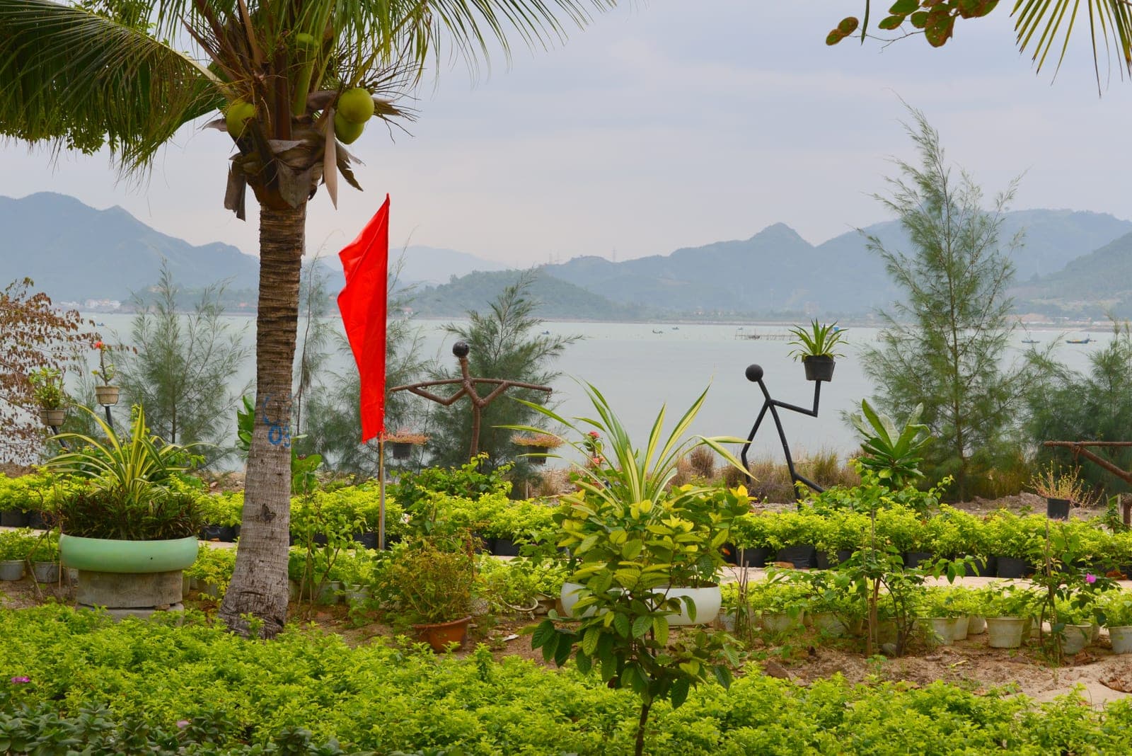 Lush waterfront garden with palm trees and red flag overlooking Nha Trang bay with mountains and fishing boats in Khanh Hoa province.